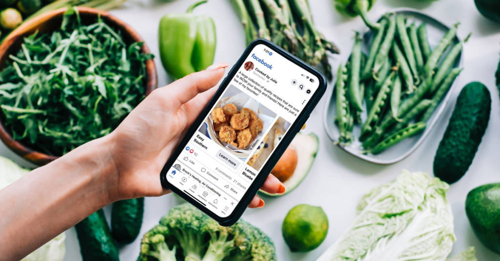 Hand holding a smartphone displaying a Facebook ad for "Cooked by Julie," surrounded by fresh green vegetables.