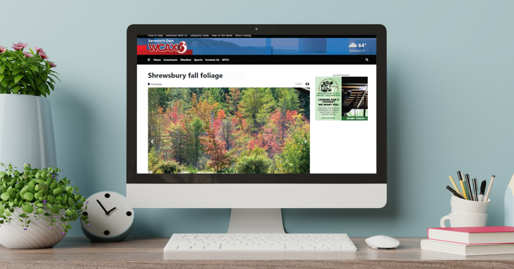 Desktop monitor displaying the WCAX3 news website with a Shrewsbury fall foliage photo gallery, styled on a desk with plants and office accessories.