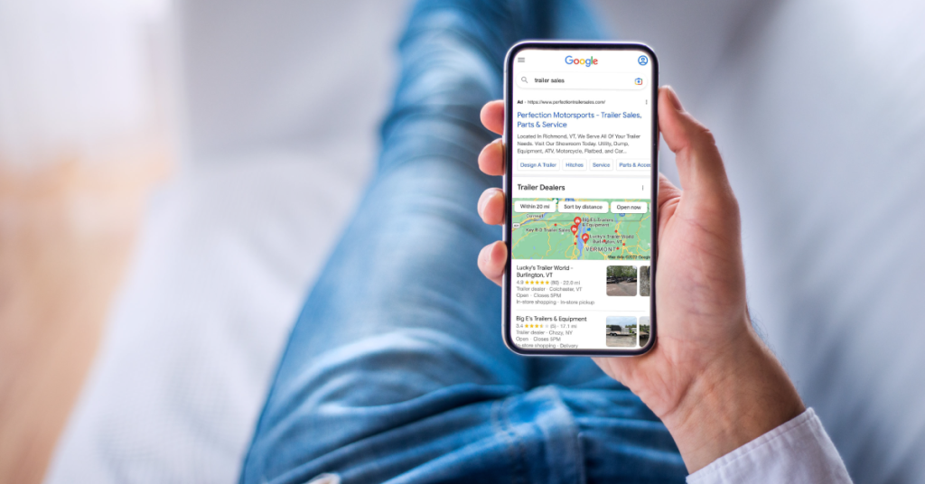 Hand holding a smartphone showing Google search results for "trailer sales" with a sponsored ad for Perfection Motorsports in Richmond, VT, and a local map pack of trailer dealers.