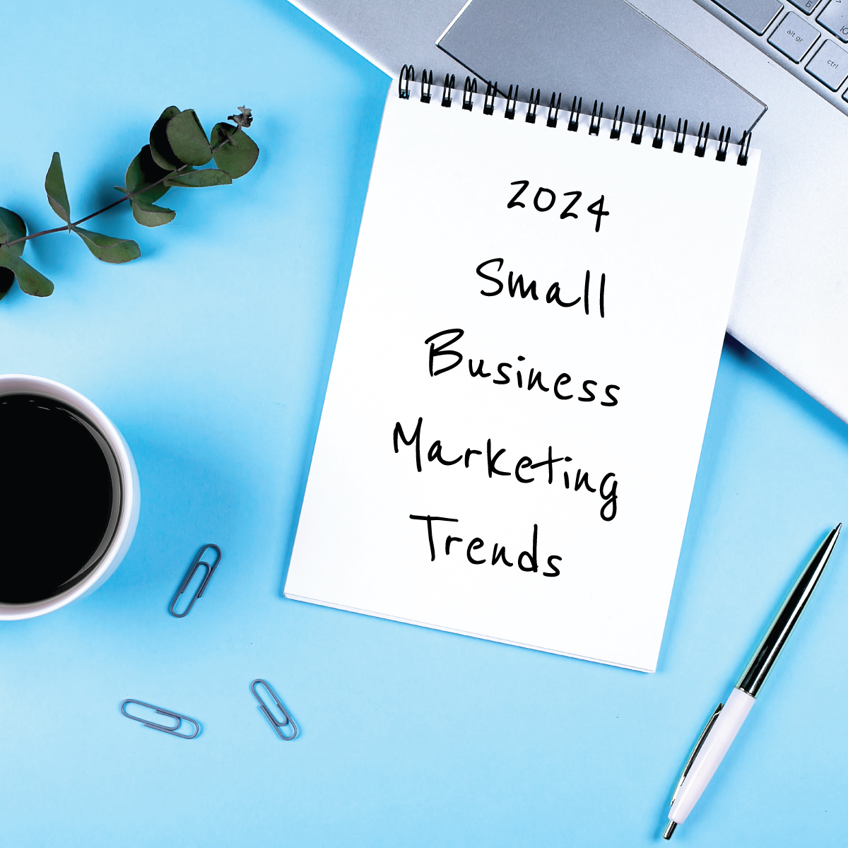 Flatlay of a notepad reading "2024 Small Business Marketing Trends" on a blue desktop with coffee, paper clips, a pen, and a laptop.