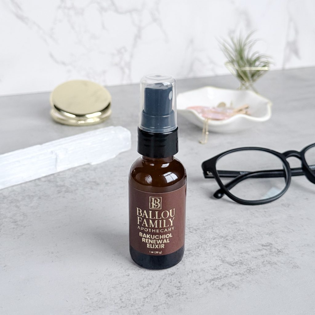 Ballou Family Apothecary Bakuchiol Renewal Elixir in an amber spray bottle, styled on a marble surface with a selenite crystal, gold compact, jewelry dish, and eyeglasses.