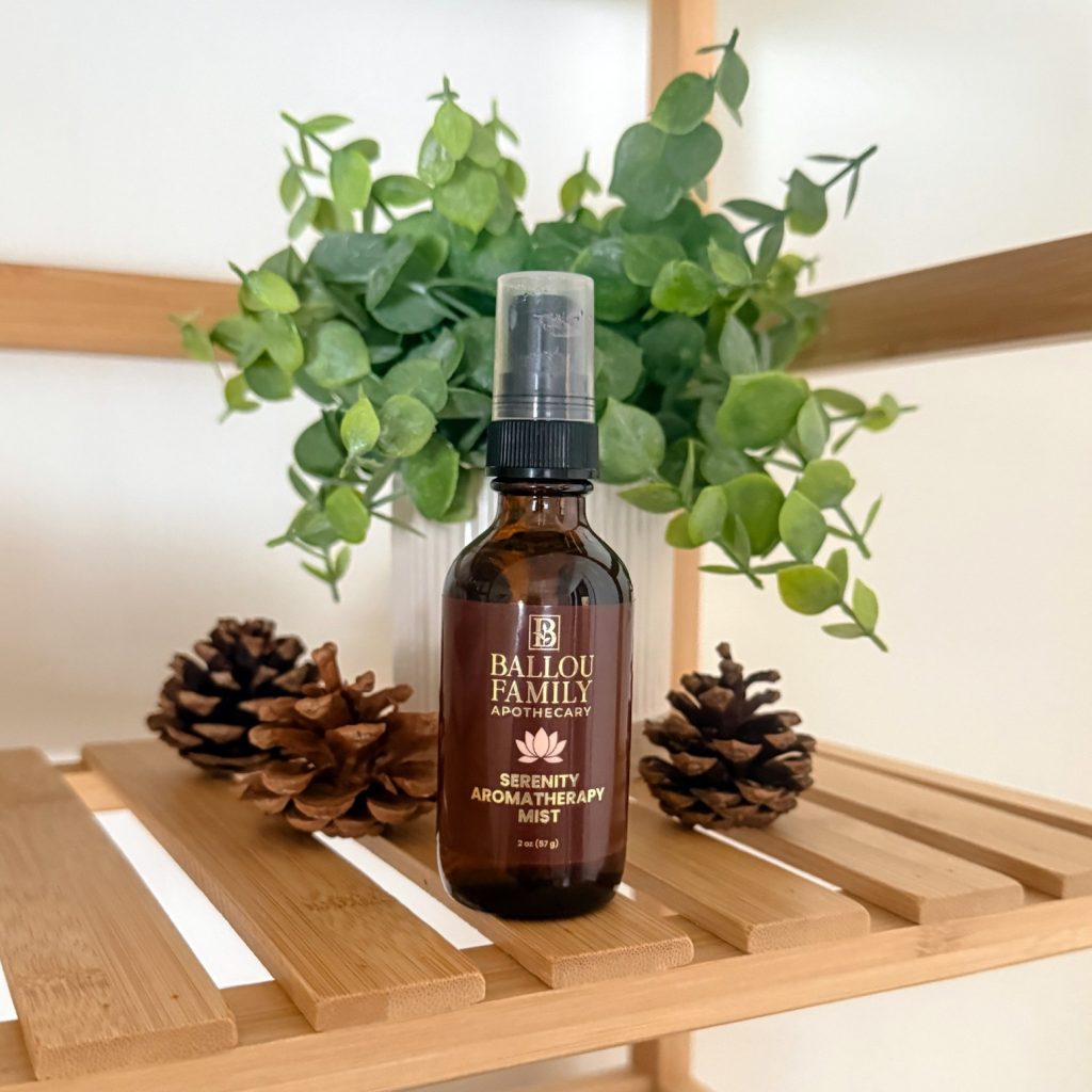 Ballou Family Apothecary Serenity Aromatherapy Mist in an amber spray bottle, styled on a bamboo shelf with pinecones and a eucalyptus plant.