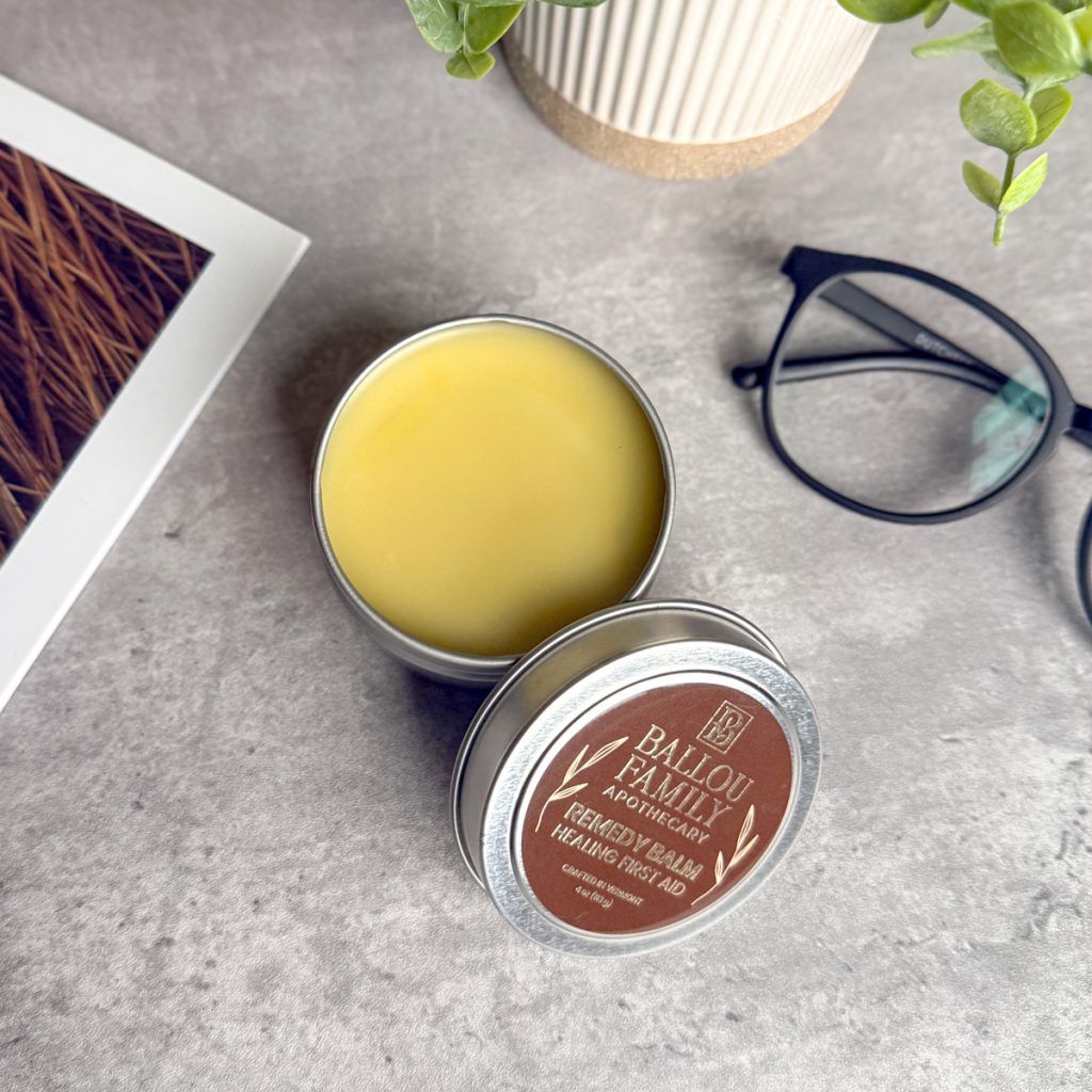 Ballou Family Apothecary Remedy Balm Healing First Aid tin, open to reveal a yellow balm, styled on a stone surface with eyeglasses and greenery.
