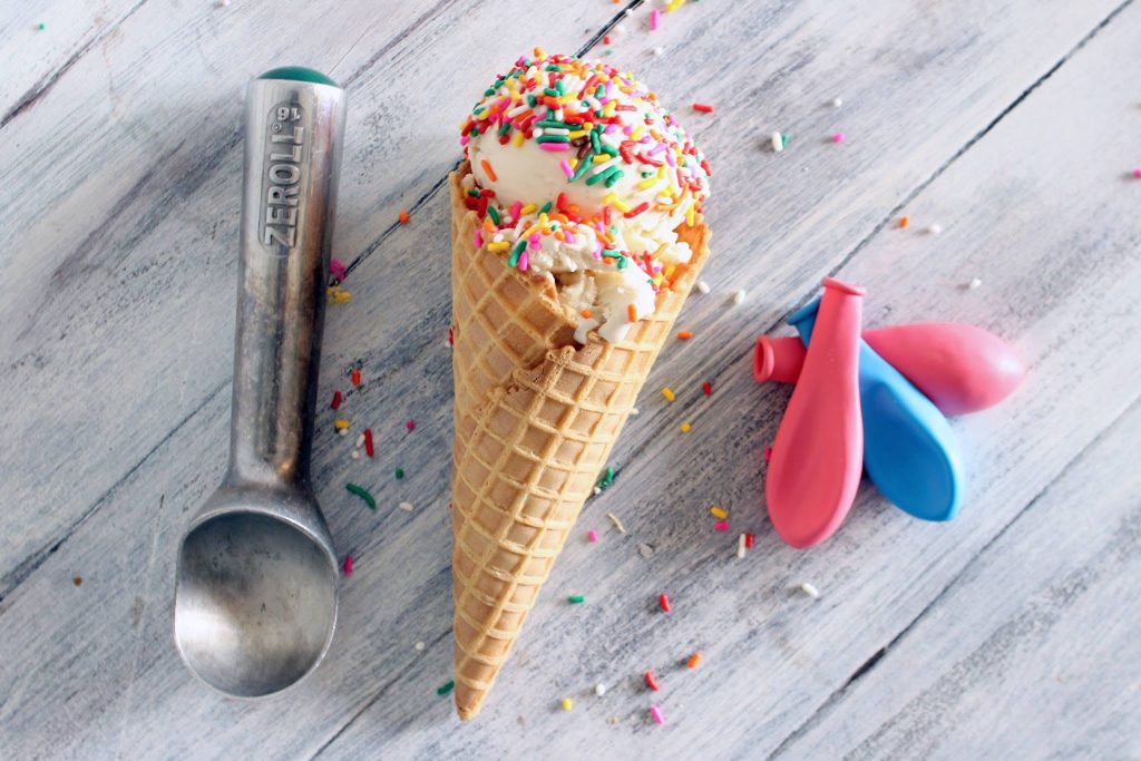 Overhead view of vanilla ice cream in a cone with rainbow sprinkles, an ice cream scoop, and balloons